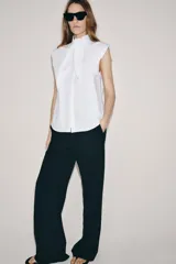 Camisa sin mangas confeccionada en algodón, con cuello redondo y detalle de lazada frontal. Presenta cierre frontal con botones ocultos por solapa y bajo redondeado.