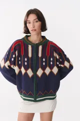 Campera tipo bomber tejida con diseño jacquard geométrico en tonos azul marino, blanco, rojo y verde. Presenta cierre frontal completo, cuello, puños y ruedo acanalados en color verde y blanco, con corte oversized y hombros caídos.