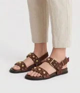 Sandalias planas Satinato de estilo sport sandal, confeccionadas en material sintético efecto gamuza color marrón. Presentan dos tiras cruzadas en el empeine decoradas con tachuelas semiesféricas doradas y ojales metálicos, además de una tira trasera con hebilla ajustable.