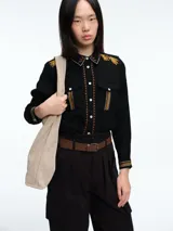 Camisa negra de manga larga con cuello clásico, cierre frontal de botones y detalles bordados en color mostaza en el cuello, hombros, tapetas de los bolsillos frontales y puños.