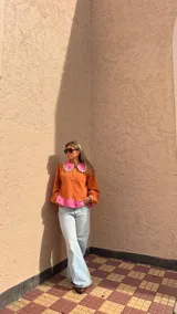 Camisa de manga larga color naranja con cuello grande en contraste color rosa y detalle de volados en el borde. Presenta un diseño con botones frontales y un detalle de tela rosa en la parte inferior.