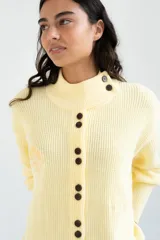 Saco tejido de punto color amarillo claro, con cuello alto asimétrico abotonado, botones oscuros contrastantes en la parte frontal y mangas largas con puños ajustados.