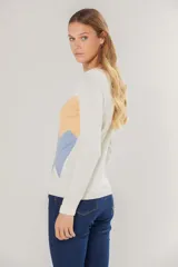 Buzo de tejido de punto fino con diseño de bloques de color en forma de zigzag en tonos lila y amarillo sobre base color crema. Presenta cuello redondo y mangas largas.