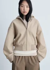 Chaqueta tipo bomber con capucha, confeccionada en tejido de poliéster con textura suave. Presenta cierre frontal de cremallera, mangas largas con puños elásticos y cintura acanalada.