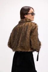 Campera corta de pelo sintético negro con detalles en cuero sintético, incluyendo bolsillos frontales con solapa, cinturón con hebilla metálica en el pecho y correas ajustables en los puños.