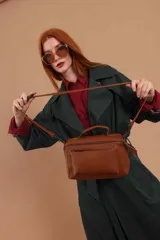 Bandolera de diseño rectangular en color suela, confeccionada en material sintético. Cuenta con un asa superior fija, correa larga ajustable y extraíble, y dos bolsillos frontales con cierre metálico, uno de ellos con diseño superpuesto. Presenta el logo de la marca grabado en el frente.