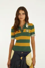 Remera polo de punto acanalado con diseño de franjas horizontales en verde y amarillo. Presenta cuello tipo polo con ribete blanco, mangas cortas con puños rayados, cierre frontal parcial con botones y parche circular bordado de la marca en el pecho.