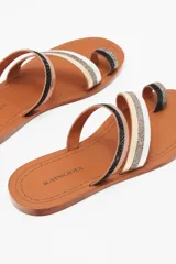 Sandalia chata de cuero vacuno con diseño de tiras finas multicolores y anillo para el dedo. Presenta tiras en color negro, dorado con acabado brillante, plateado metalizado y beige sobre una suela plana de cuero marrón.