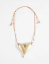 Collar con cordón ajustable de color beige claro y un dije grande con forma de corazón tridimensional, acabado en tono dorado envejecido o plateado antiguo.