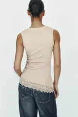 Camiseta ajustada (slim fit) color crema, con cuello redondo y mangas sisa. Presenta frunces laterales y un bajo asimétrico con detalle de encaje a tono.