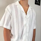 Camisa de tejido calado con textura, color blanco, de corte oversize, manga corta y cuello solapa con cierre frontal de botones.