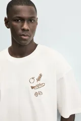 Camiseta de manga corta color blanco roto, corte relajado, con un pequeño bordado/estampado en el pecho que incluye una manzana, una rama de laurel, una trompeta y dos dados.