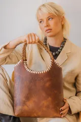 Bolso tipo matera de cuero con textura de reptil, color tostado, con manijas trenzadas y detalles de costura artesanal en los bordes.