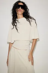 Top fluido de color beige con cuello perkins y mangas acampanadas por debajo del codo. Presenta un diseño de bajo redondeado y textura sutil en la tela.