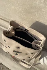 Bandolera de cuero color beige con diseño de ojales metálicos distribuidos por toda la superficie frontal y placa metálica con logo de la marca. Cuenta con correa larga ajustable.