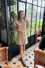 Vestido corto de lino color beige, con cuello alto, mangas cortas abullonadas con puños elásticos y volados en el pecho. Cuenta con cierre frontal de botones.