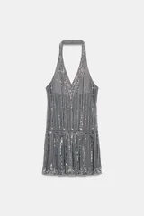 Vestido corto gris, estilo flapper o años 20, con cuello halter y escote pronunciado en pico. Confeccionado en tul con apliques verticales de lentejuelas y brillos plateados. Presenta un ruedo con volado.