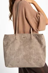Bolso tipo tote de gran tamaño confeccionado en gamuza sintética color tostado. Presenta un diseño minimalista con asas largas de hombro y una pequeña placa metálica frontal con el logo de la marca.