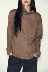 Top de cuello alto y manga larga, confeccionado en tejido de algodón gris melange. Presenta un diseño con frunces laterales ajustados mediante un aplique metálico decorativo.