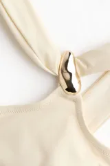 Top de bikini asimétrico de un solo hombro en color beige, con doble tirante y un detalle de aplique metálico dorado en la parte delantera.