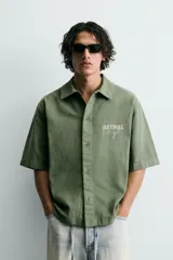 Camisa de corte relajado color verde oliva, con cuello solapa, manga corta y cierre frontal de botones. Presenta un estampado con el texto 'ASTRAL Drift' en el frente.