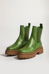 Bota tipo chelsea con suela track de goma en color marrón y capellada de cuero negro. Presenta elásticos laterales y tirador trasero.