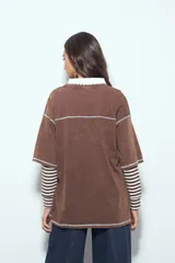 Remera de manga corta color marrón con efecto desgastado, corte oversize y costuras a la vista en color blanco. Presenta cuello redondo con cuello polo blanco superpuesto y mangas largas a rayas horizontales marrones y blancas por debajo.