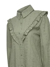 Blusa verde de algodón orgánico con cuello camisero, manga larga y cierre frontal de botones. Presenta volantes bordados que recorren el frente, la espalda y las sisas, además de detalles bordados en toda la prenda.