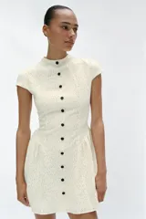Vestido corto blanco de encaje, con cuello redondo, manga corta y bajo evasé con frunces. Cuenta con fila de falsos botones negros delanteros a contraste y cierre trasero con cremallera oculta.