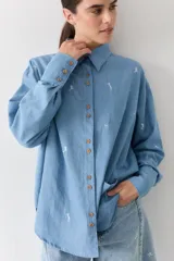 Camisa de jean azul claro con bordados pequeños en color blanco de figuras deportivas. Presenta cuello clásico, cierre frontal con botones de madera y mangas largas con puños abotonados.
