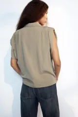 Camisa fluida de cuello solapa y manga corta, con diseño a rayas en tonos beige y blanco, detalle de pliegues en los hombros y cierre frontal con botones.