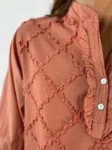 Blusa color beige con cuello mao, mangas 3/4 con puños elásticos y detalle de diseño geométrico texturizado en el frente.
