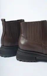 Bota corta tipo Chelsea de cuero negro, con puntera cuadrada y suela track gruesa y dentada. Presenta paneles elásticos acanalados a los lados para facilitar el calce.