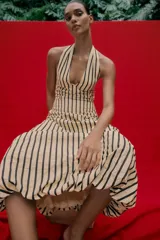 Vestido midi de corte globo con estampado de rayas verticales, cuello halter anudado, escote en pico y espalda descubierta. Presenta bajo abullonado con elástico interior y cierre lateral con cremallera oculta.