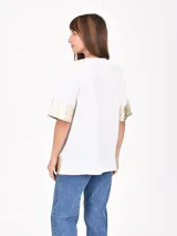 Remera blanca de corte holgado con detalles metalizados en color dorado en las mangas y el ruedo. Presenta un estampado minimalista con texto en el frente.
