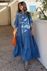 Vestido largo de estilo camisero en color azul, con cuello clásico, cierre frontal de botones y mangas tres cuartos con trabillas. Presenta un diseño de falda con cortes horizontales que le otorgan volumen y movimiento.