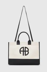 Tote bag de diseño estructurado, confeccionado en lona color crema con base, asas y detalles en cuero negro. Presenta un logo circular en relieve en el frente y correa larga ajustable para llevar al hombro.