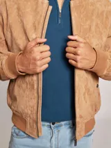 Campera bomber color marrón claro confeccionada en gamuza, con cierre frontal y bolsillos laterales con botón a presión. Presenta cuello, puños y cintura acanalados.