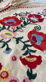 Chaleco de algodón crudo sin mangas, con escote en V y cierre frontal con tiras para atar. Presenta un diseño corto con bordados florales coloridos en tonos rojos, azules y amarillos, y detalles geométricos rojos en los bordes.
