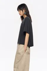 Camiseta de manga corta y escote redondo, color beige claro, con corte boxy. Presenta una hilera de tachuelas metálicas negras decorativas en la parte superior del pecho y en las costuras de los hombros.