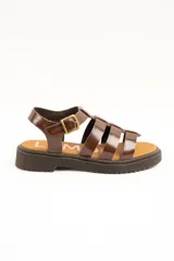 Sandalias cangrejeras de cuero marrón charol, estilo romano con múltiples tiras cruzadas, suela track plana y cierre de hebilla metálica ajustable en el tobillo.