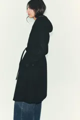 Abrigo de paño negro con capucha, cierre frontal cruzado con botones y cinturón de lazada en el mismo tejido. Presenta manga larga con trabillas en los puños y bolsillos laterales.