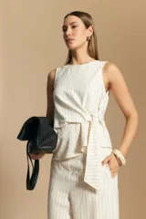 Top sin mangas de color blanco hueso con finas rayas verticales oscuras. Presenta un diseño con un cruce lateral que forma una lazada o nudo en la cintura.