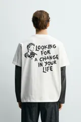 Camiseta blanca de corte relajado, con cuello redondo y manga corta. Presenta un pequeño estampado de texto en contraste en la parte frontal izquierda del pecho.