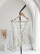 Blusa o cárdigan corto tejido en crochet o macramé color blanco, con diseño de ondas o chevron y flecos en el ruedo. Presenta un escote en V y se ata al frente con un lazo.