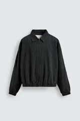 Cazadora de hombre de ajuste cropped fit, confeccionada en tejido de algodón con patrón de rayas verticales finas en tonos oscuros (negro/azul marino y marrón/beige). Presenta cuello solapa, cierre frontal de cremallera, bolsillos laterales y elástico en puños y bajo.