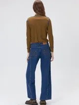 Jeans de corte ancho (wide leg) y tiro medio, confeccionados en denim de algodón negro con costuras contrastantes en color ocre. Presenta múltiples botones metálicos frontales y tachas metálicas aplicadas a lo largo de los laterales y la pretina.