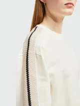 Sudadera color crema de corte holgado con cuello redondo y mangas largas. Presenta una banda lateral con diseño de tejido calado y logo de la marca en la manga izquierda.