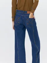Jeans de corte ancho (wide leg) y tiro medio, confeccionados en denim de algodón negro con costuras contrastantes en color ocre. Presenta múltiples botones metálicos frontales y tachas metálicas aplicadas a lo largo de los laterales y la pretina.