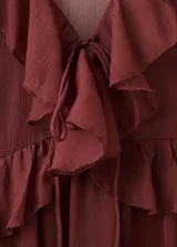 Vestido largo de tejido fluido color bordó, de diseño evasé con escote en pico y mangas largas con puños elásticos. Presenta detalles de volantes en el cuello y forro interior.